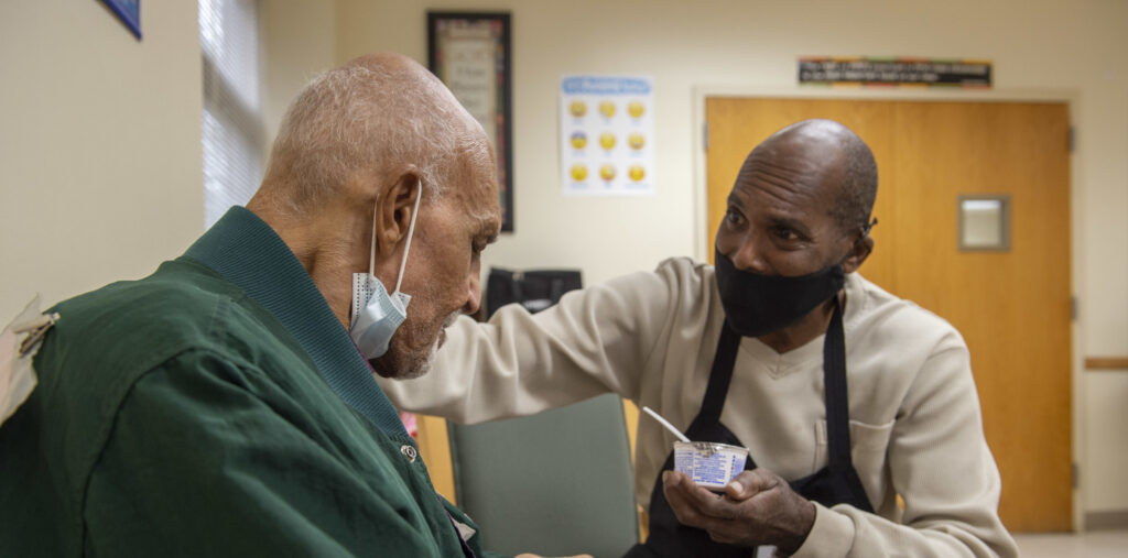 Catholic Charities person holding a yogurt container with a spoon helping and older man